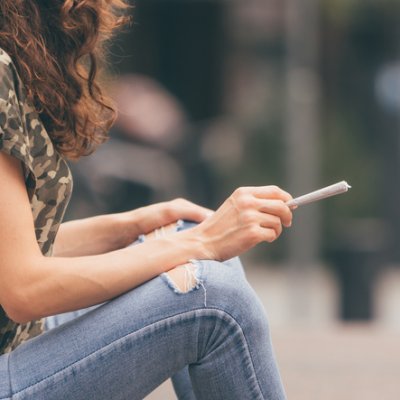 Image of a woman wearing jeans sitting down with a joint in her hands 